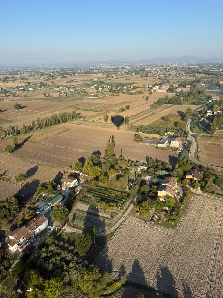 Veduta aerea della Valle Umbra al mattino, con l'ombra della mongolfiera sui campi e Assisi in lontananza